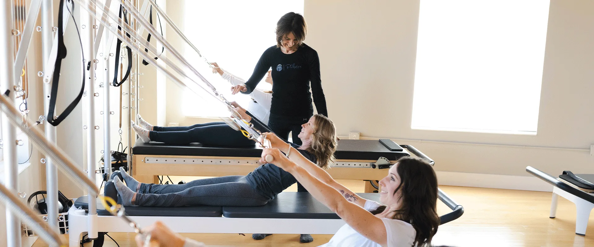 A Bovée Pilates instructor guides two clients on reformers with a tower in the bright, light-wood studio.